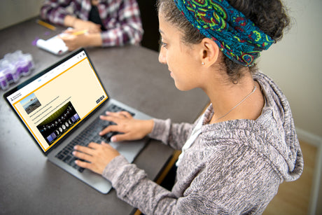 Person using a laptop with another person in the background at a table.